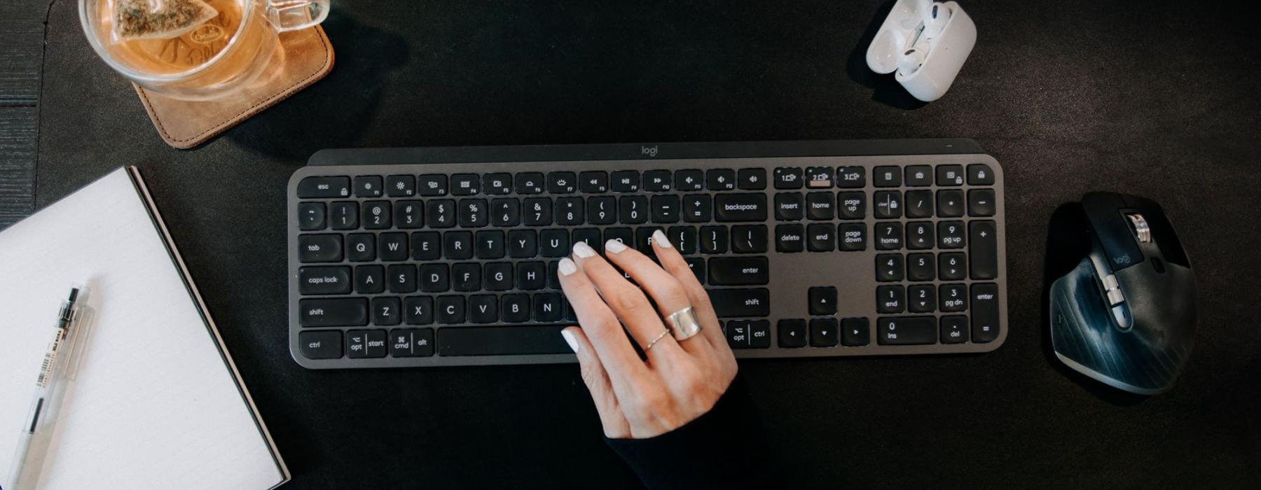 woman's hand on a keyboard surrounded by office items and a cup of tea on a coaster
